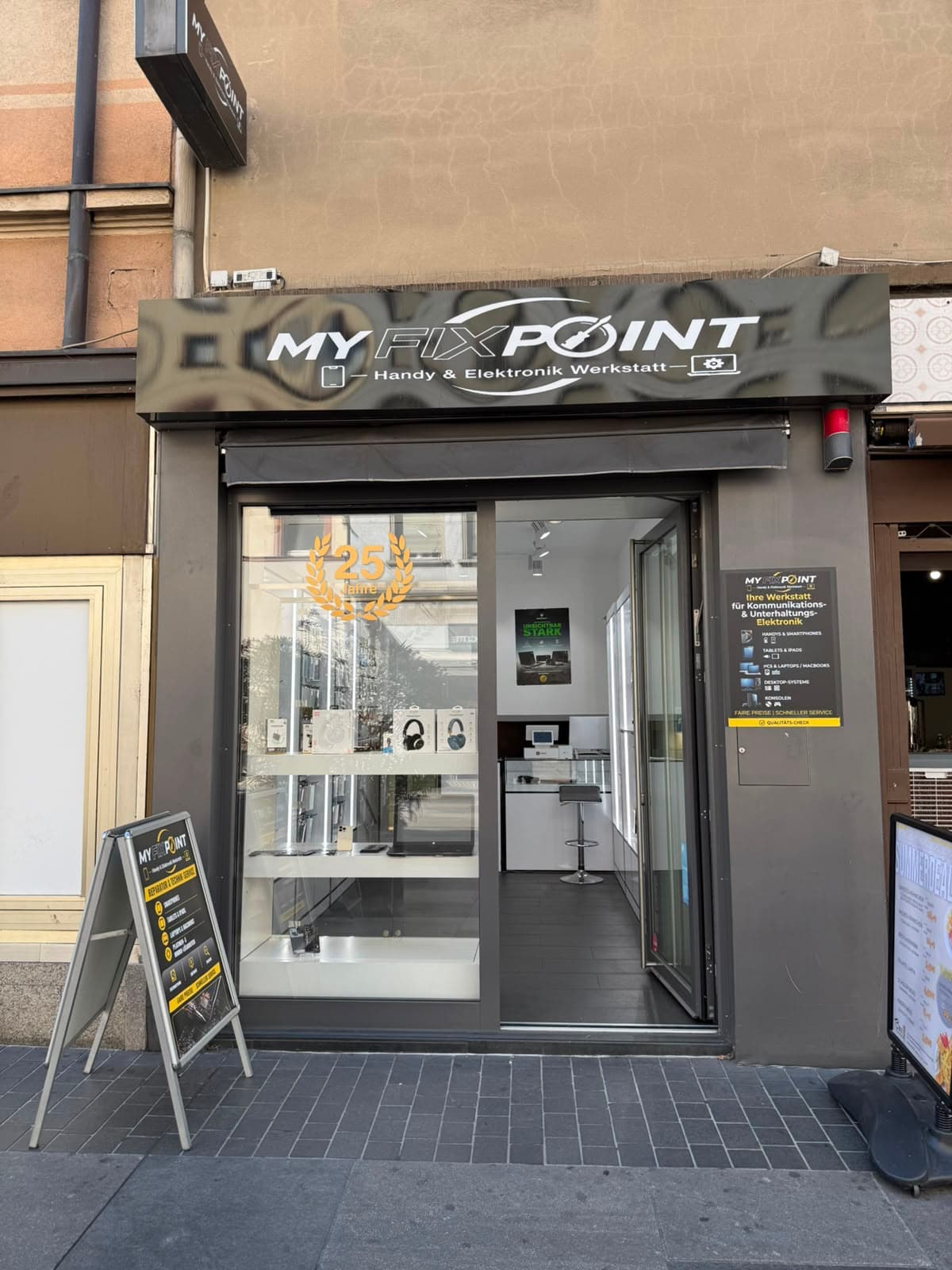 Ladenfront von MY FIX POINT bei Tag, mit beleuchtetem Schild "Handy & Elektronik Werkstatt" und offener Eingangstür.